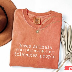 May include: A coral-colored t-shirt with the text "loves animals tolerates people" in white. The shirt has a small white tag with the text "COMFORT COLORS" on it. There are small white animal and flower graphics below the text.