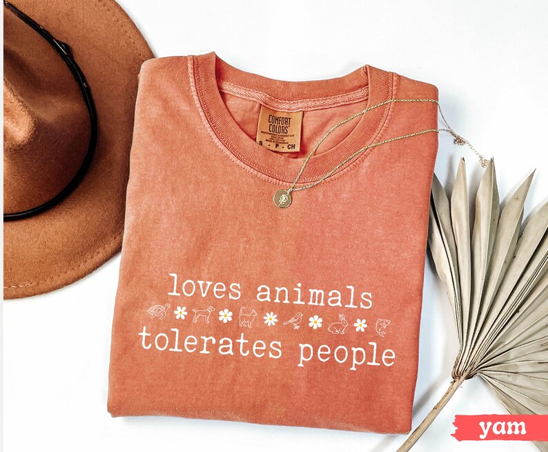 May include: A coral-colored t-shirt with the text "loves animals tolerates people" in white. The shirt has a small white tag with the text "COMFORT COLORS" on it. There are small white animal and flower graphics below the text.