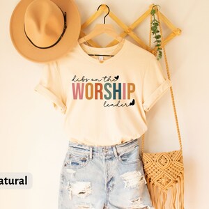 Puede incluir: Una camiseta beige con el texto "dibs on the WORSHIP leader" en colores arco&iacute;ris. La camiseta est&aacute; colgada en un estante de madera con un sombrero marr&oacute;n y una bolsa tejida.