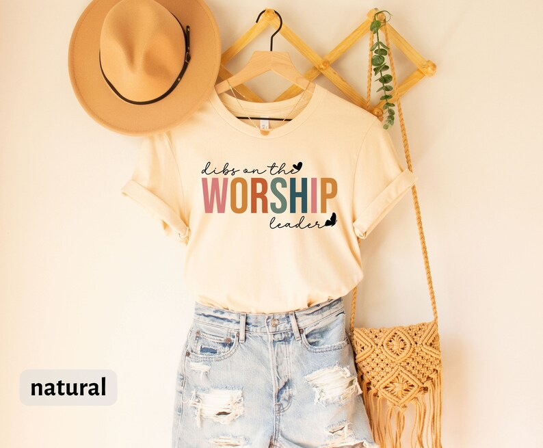Puede incluir: Una camiseta beige con el texto "dibs on the WORSHIP leader" en colores arco&iacute;ris. La camiseta est&aacute; colgada en un estante de madera con un sombrero marr&oacute;n y una bolsa tejida.