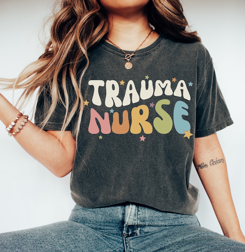 May include: A dark grey t-shirt with the text "TRAUMA NURSE" in colorful, retro-style lettering. The text is surrounded by small, white stars.