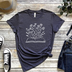 K&ouml;nnte beinhalten: Ein dunkelgraues T-Shirt mit einem wei&szlig;en Umriss eines Buches, aus dem Blumen wachsen.