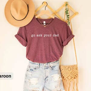 May include: A maroon t-shirt with the text "go ask your dad" in white lettering. The shirt is hanging on a wooden hanger and is paired with denim shorts and a straw bag.