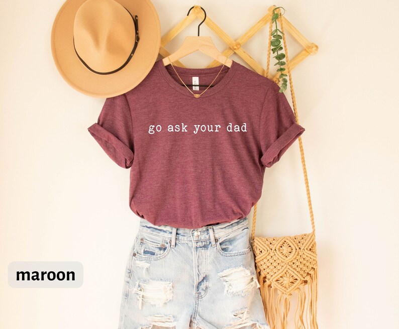 May include: A maroon t-shirt with the text "go ask your dad" in white lettering. The shirt is hanging on a wooden hanger and is paired with denim shorts and a straw bag.