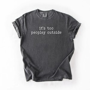 May include: A dark gray t-shirt with the text "it's too peopley outside" printed in white.