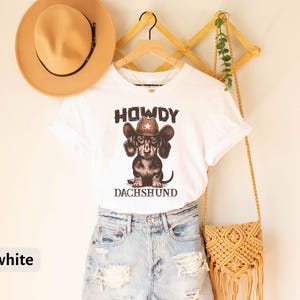 Puede incluir: Camiseta blanca con un dise&ntilde;o de perro salchicha con sombrero de vaquero, con las palabras "HOWDY" y "DACHSHUND". La camiseta se combina con pantalones cortos de mezclilla y un sombrero beige. Una bolsa de macram&eacute; cuelga cerca.