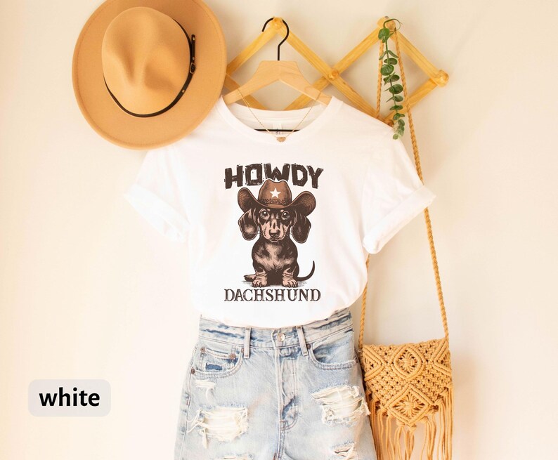 Puede incluir: Camiseta blanca con un dise&ntilde;o de perro salchicha con sombrero de vaquero, con las palabras "HOWDY" y "DACHSHUND". La camiseta se combina con pantalones cortos de mezclilla y un sombrero beige. Una bolsa de macram&eacute; cuelga cerca.