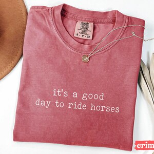 May include: A crimson red t-shirt with the text "it's a good day to ride horses" in white. The shirt is made of a soft, comfortable material and has a relaxed fit.