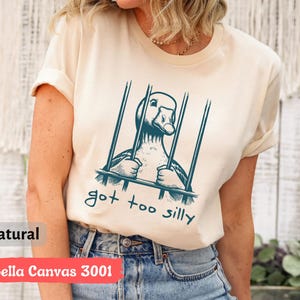 May include: A beige t-shirt with a graphic of a goose in a cage and the text "got too silly".