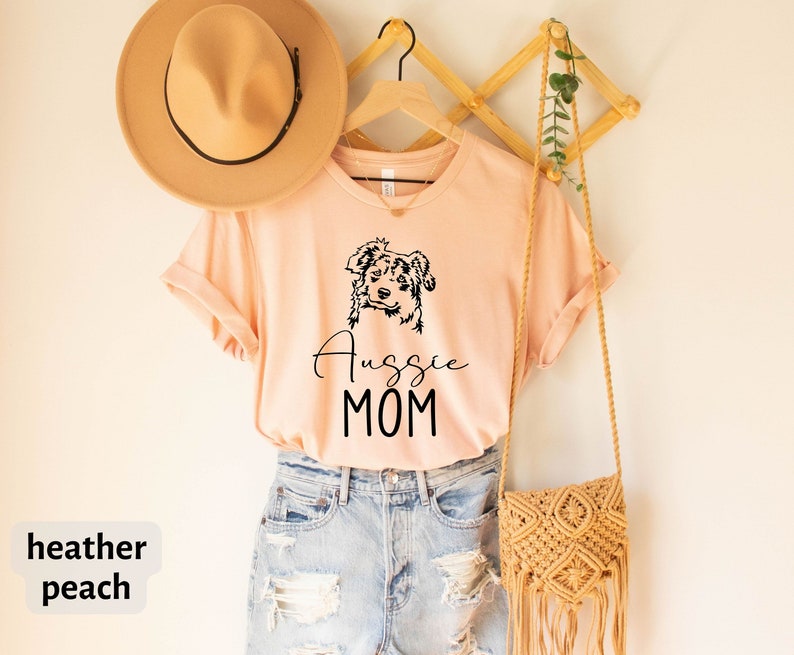 K&ouml;nnte beinhalten: Ein pfirsichfarbenes T-Shirt mit einer Schwarzwei&szlig;illustration eines Australian Shepherds und dem Text "Aussie Mom".