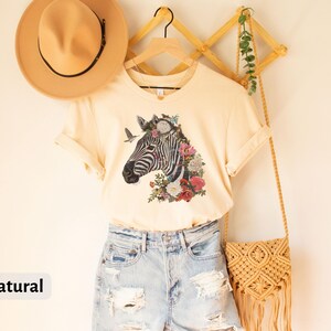 May include: A natural colored t-shirt with a floral zebra graphic. The shirt is hanging on a wooden rack with a straw bag and a hat.