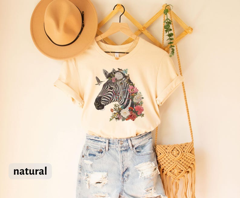 May include: A natural colored t-shirt with a floral zebra graphic. The shirt is hanging on a wooden rack with a straw bag and a hat.
