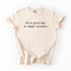 May include: A beige t-shirt with the text "it's a good day to make cookies" printed in black.