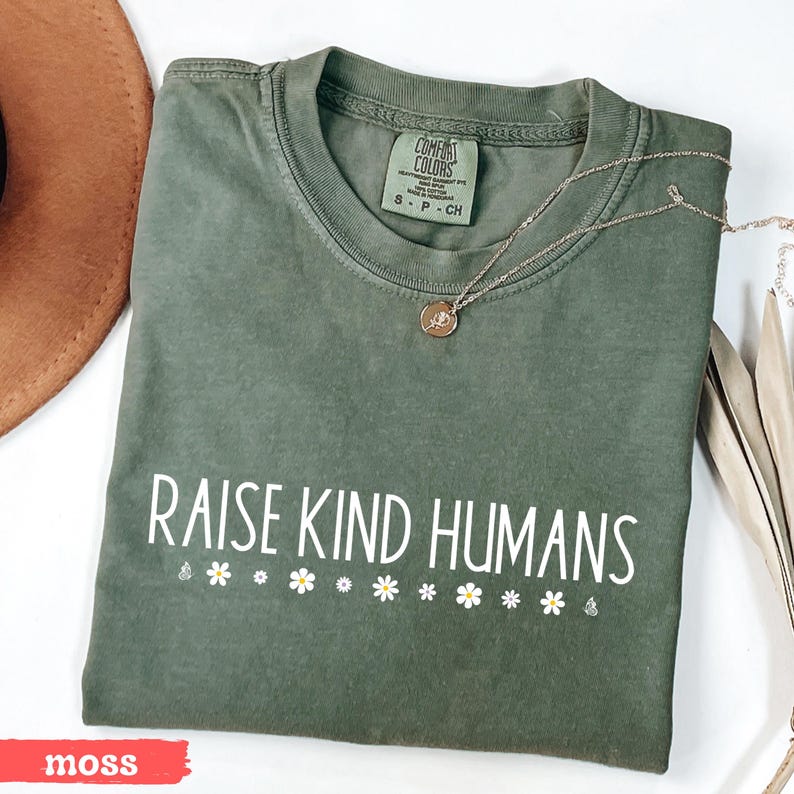 Op de afbeelding: Een groen T-shirt met de tekst "Raise Kind Humans" in witte letters met een rij witte madeliefjes eronder. Het shirt is gemaakt van een zacht, comfortabel materiaal.