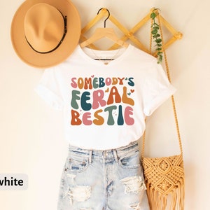 May include: White t-shirt with the text "Somebody's Feral Bestie" in colorful, retro-style lettering.