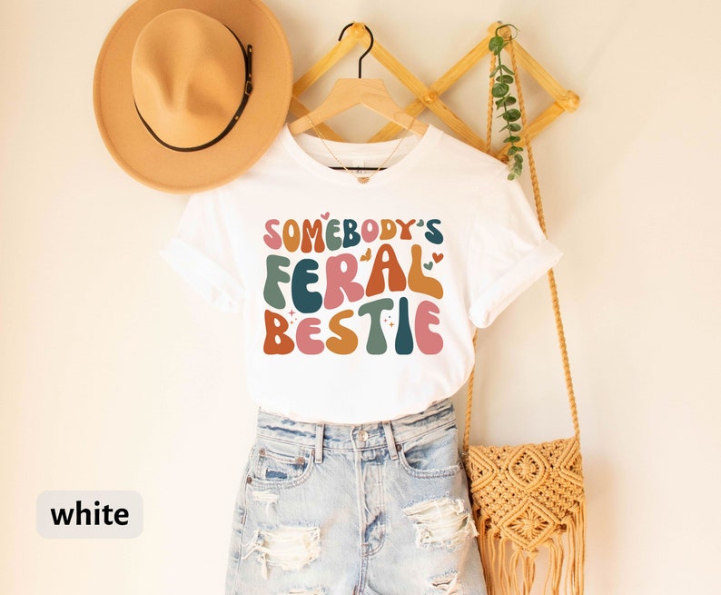 May include: White t-shirt with the text "Somebody's Feral Bestie" in colorful, retro-style lettering.