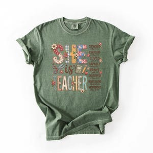 May include: A green t-shirt with rolled-up sleeves. The shirt features the text "SHE IS TEACHER" in a colorful, floral design. Additional words like "STRONG", "CHOSEN", and "BRAVE" are also included in the design.