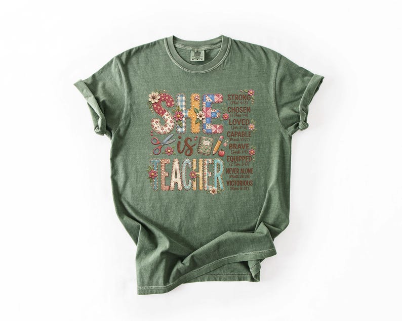 May include: A green t-shirt with rolled-up sleeves. The shirt features the text "SHE IS TEACHER" in a colorful, floral design. Additional words like "STRONG", "CHOSEN", and "BRAVE" are also included in the design.