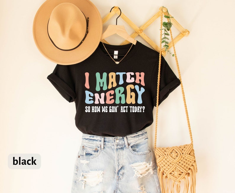 May include: Black t-shirt with the text "I Match Energy So How We Gon' Act Today?" in colorful letters. The shirt is hanging on a wooden rack with a brown hat and a woven bag.