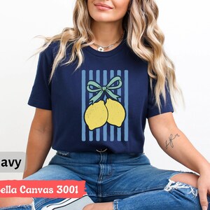May include: A navy blue t-shirt with a graphic of two lemons tied together with a blue bow on a blue and white striped background. The text "Bella Canvas 3001" is printed on the bottom of the shirt.