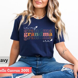 May include: A navy blue t-shirt with the text "in my grandma era" in rainbow colors. The shirt is made of Bella Canvas 3001 fabric.
