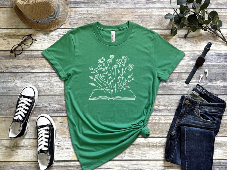 K&ouml;nnte beinhalten: Ein gr&uuml;nes T-Shirt mit einer wei&szlig;en Grafik eines offenen Buches, aus dem Blumen wachsen.