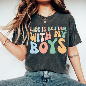 K&ouml;nnte beinhalten: Ein dunkelgraues T-Shirt mit dem Text "Life is better with my boys" in bunten, stilisierten Buchstaben.