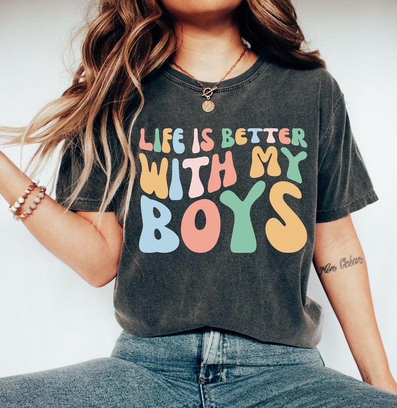 K&ouml;nnte beinhalten: Ein dunkelgraues T-Shirt mit dem Text "Life is better with my boys" in bunten, stilisierten Buchstaben.