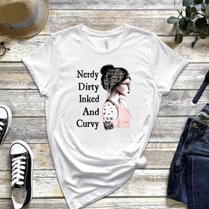 Op de afbeelding: Wit T-shirt met een afbeelding van een vrouw met boeken als haar en een tatoeage op haar arm. De tekst op het shirt luidt "Nerdy Dirty Inked And Curvy".