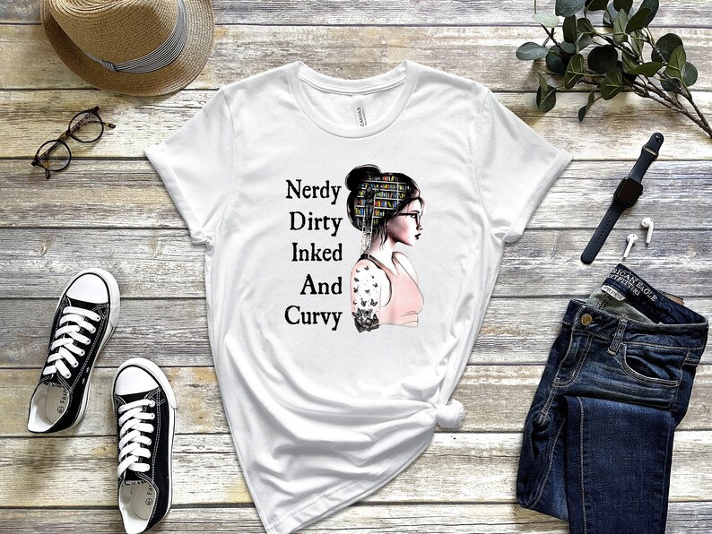 Op de afbeelding: Wit T-shirt met een afbeelding van een vrouw met boeken als haar en een tatoeage op haar arm. De tekst op het shirt luidt "Nerdy Dirty Inked And Curvy".