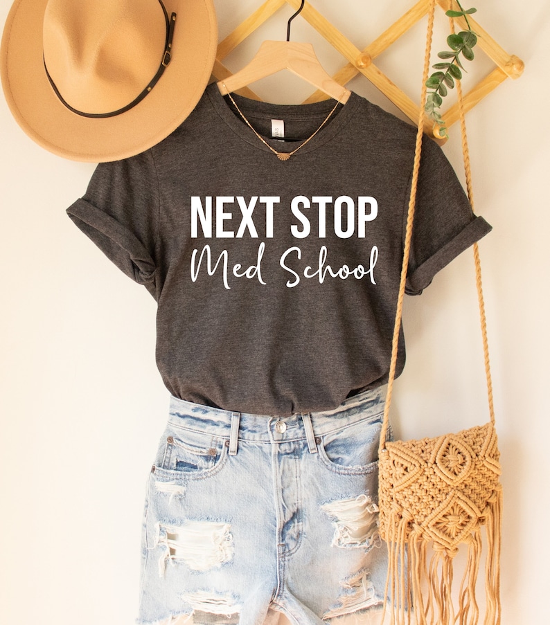 May include: A dark gray t-shirt with the text "NEXT STOP Med School" in white. The shirt is hanging on a wooden hanger and is styled with denim shorts and a woven bag.