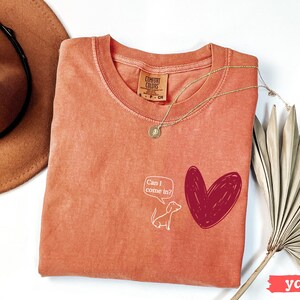 May include: A yam-colored Comfort Colors t-shirt with a round neckline. The shirt features a white cartoon dog and a red heart graphic. Text in a speech bubble reads "Can I come in?" A gold necklace and a brown hat are also visible.