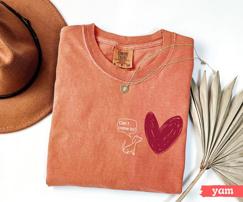 May include: A yam-colored Comfort Colors t-shirt with a round neckline. The shirt features a white cartoon dog and a red heart graphic. Text in a speech bubble reads "Can I come in?" A gold necklace and a brown hat are also visible.