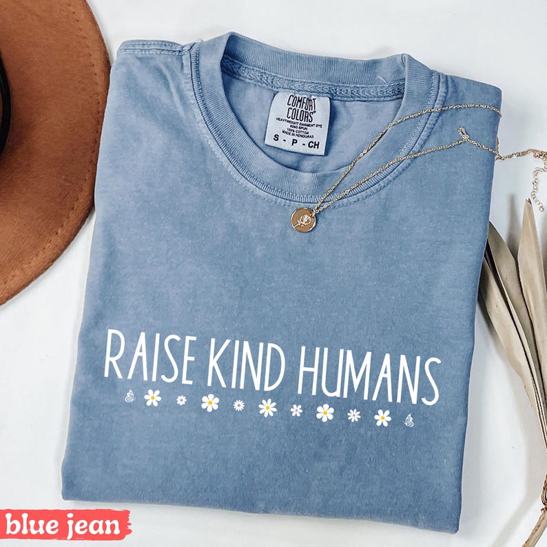 Op de afbeelding: Een jeansblauw T-shirt met de tekst "Raise Kind Humans" in witte letters en een rij witte madeliefjes onder de tekst.
