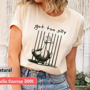 May include: A white t-shirt with a black and white illustration of a goose in a cage. The text "got too silly" is printed on the shirt.