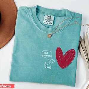 May include: Seafoam green t-shirt with a white cartoon dog and a red heart graphic. The dog has a speech bubble that says "Can I come in?" A gold necklace with a pendant is draped across the shirt.