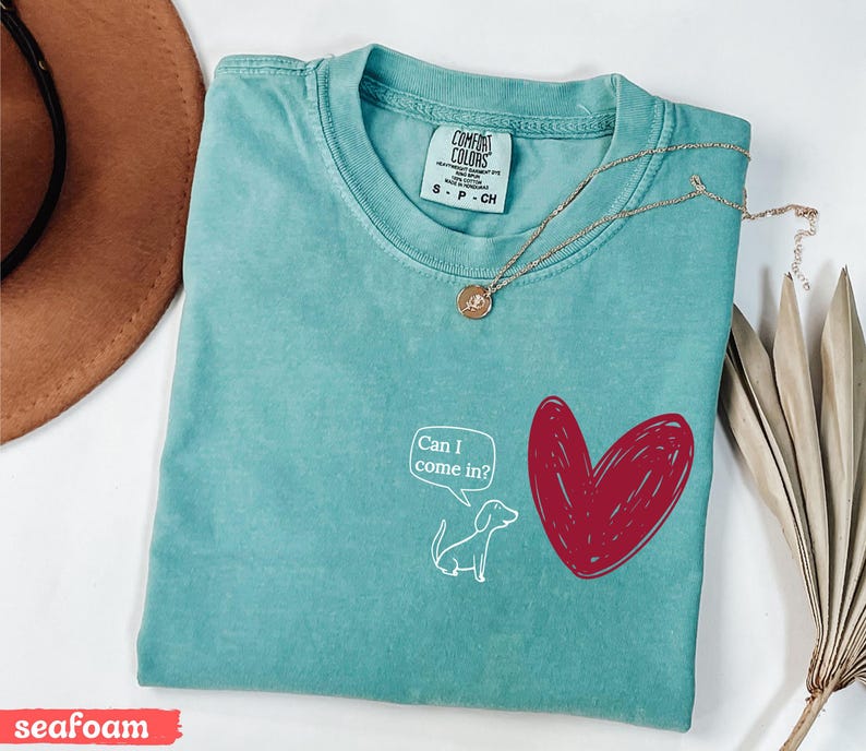 May include: Seafoam green t-shirt with a white cartoon dog and a red heart graphic. The dog has a speech bubble that says "Can I come in?" A gold necklace with a pendant is draped across the shirt.