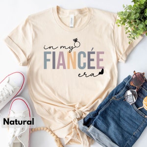 May include: A beige t-shirt with the text "in my fianc&eacute;e era" in colorful letters with a diamond ring graphic above the text. A butterfly graphic is to the right of the text.