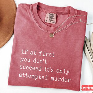 Op de afbeelding: Karmizijnrood T-shirt met de witte tekst "if at first you don't succeed it's only attempted murder". Een zilveren ketting met hanger ligt op het shirt. Het shirt heeft een ronde hals en korte mouwen.