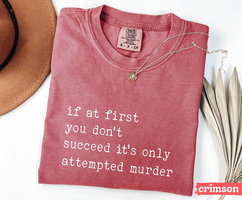 Op de afbeelding: Karmizijnrood T-shirt met de witte tekst "if at first you don't succeed it's only attempted murder". Een zilveren ketting met hanger ligt op het shirt. Het shirt heeft een ronde hals en korte mouwen.