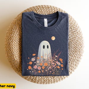 Pu&ograve; includere: T-shirt blu navy melange con un design a tema fantasma. Il fantasma bianco con occhi neri &egrave; in un campo di fiori arancioni e rosa sotto una luna piena. La t-shirt &egrave; presentata su una superficie intrecciata.