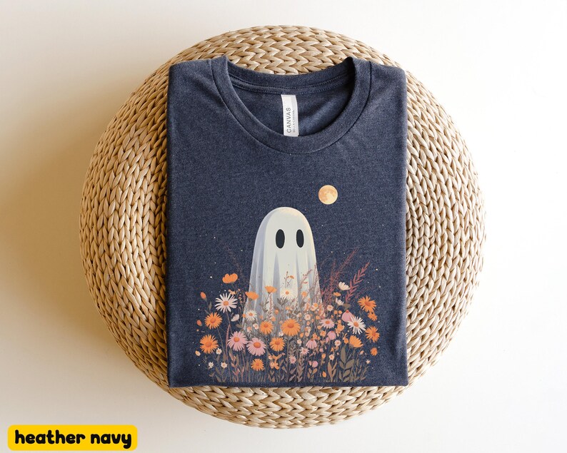 Pu&ograve; includere: T-shirt blu navy melange con un design a tema fantasma. Il fantasma bianco con occhi neri &egrave; in un campo di fiori arancioni e rosa sotto una luna piena. La t-shirt &egrave; presentata su una superficie intrecciata.