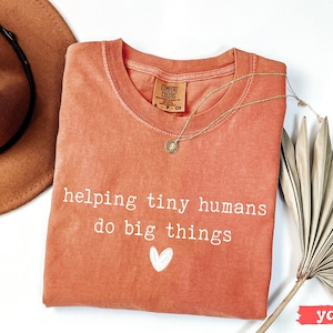 May include: A coral-colored Comfort Colors t-shirt with the text "helping tiny humans do big things" and a white heart. A gold necklace and a brown hat are also in the image. A dried palm leaf is on the right.