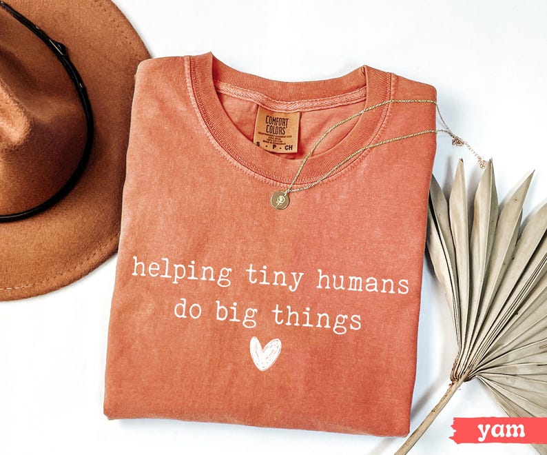 May include: A coral-colored Comfort Colors t-shirt with the text "helping tiny humans do big things" and a white heart. A gold necklace and a brown hat are also in the image. A dried palm leaf is on the right.