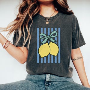May include: A dark gray t-shirt with a graphic of two yellow lemons tied together with a blue bow on a blue and white striped background.