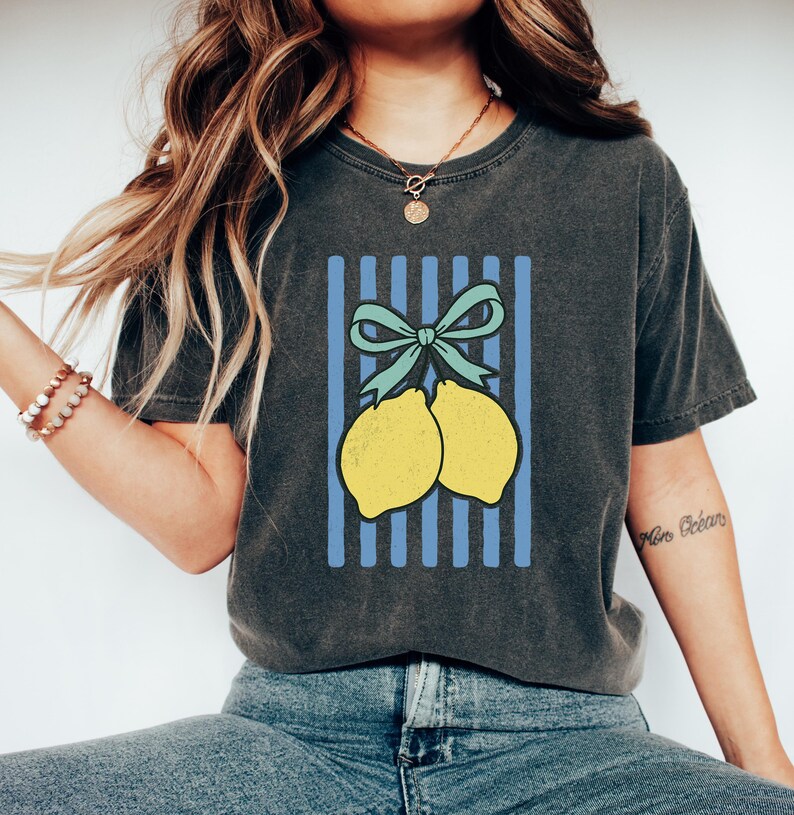 May include: A dark gray t-shirt with a graphic of two yellow lemons tied together with a blue bow on a blue and white striped background.