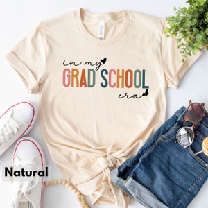 May include: A beige t-shirt with the text "in my grad school era" in colorful letters and a small black heart. The shirt is laid out on a white surface with a pair of white sneakers, a pair of blue denim shorts, and a wooden bead necklace.