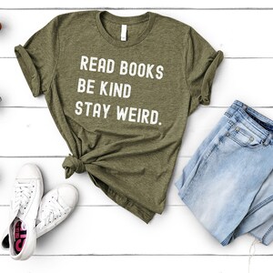 May include: A green t-shirt with white text that reads "Read Books Be Kind Stay Weird." The shirt is laid out on a white wooden surface with a pair of white sneakers, a pair of blue jeans, and three small potted plants.