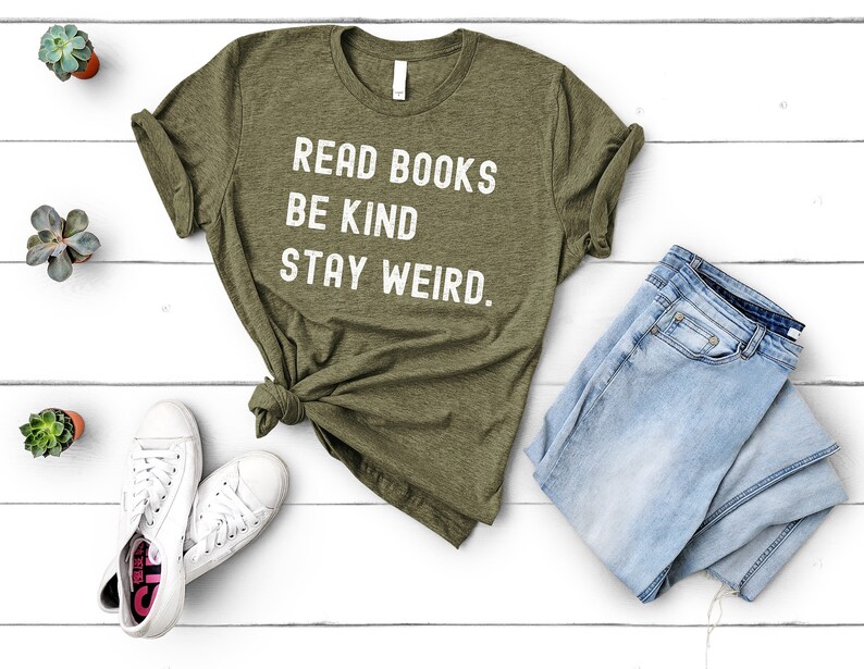 May include: A green t-shirt with white text that reads "Read Books Be Kind Stay Weird." The shirt is laid out on a white wooden surface with a pair of white sneakers, a pair of blue jeans, and three small potted plants.