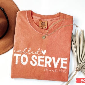 Pu&ograve; includere: Una maglietta Comfort Colors color igname con le parole "called TO SERVE" in bianco, insieme al versetto biblico Marco 10:45. Una collana d'oro e un cappello marrone sono anche nell'immagine.